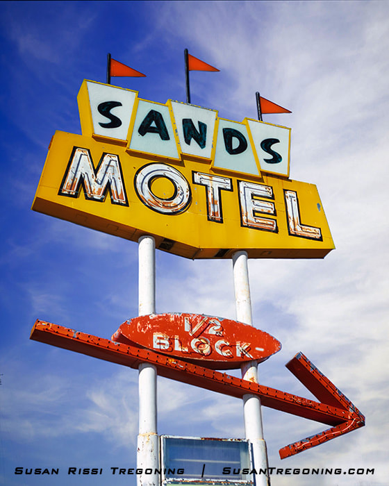A vintage roadside sign with yellow and blue panels reading “Sands Motel” stands against a blue sky. A red arrow below points to the right with the words “½ Block.” The sign is mounted on two white poles.