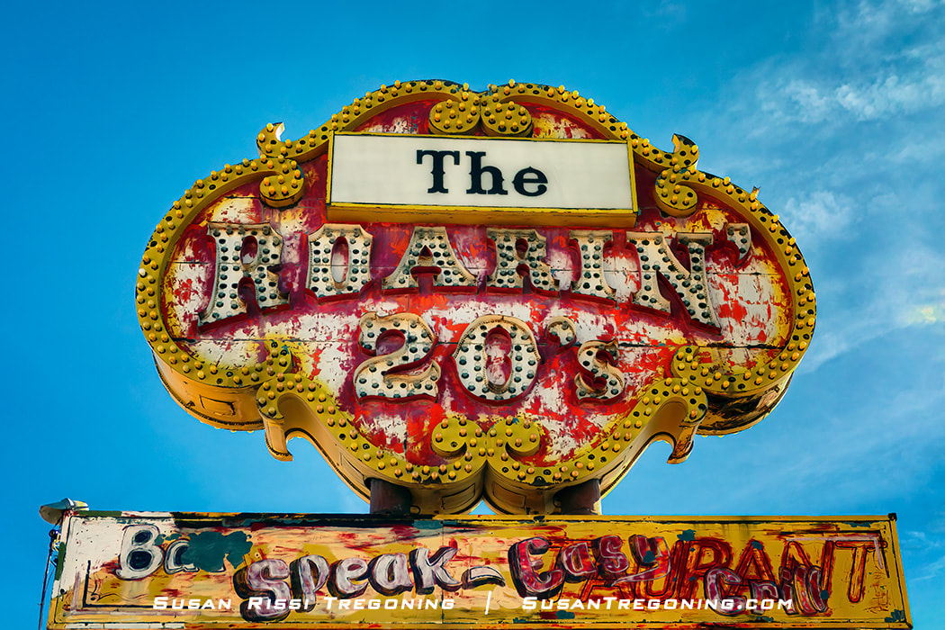 A vintage roadside sign with bold lettering reading “The Roarin’ 20s” stands against a clear blue sky. The sign has an ornate yellow border with decorative scrollwork and a smaller panel below with the words “Be Speak‑Easy Restaurant.” The paint is faded and chipped.