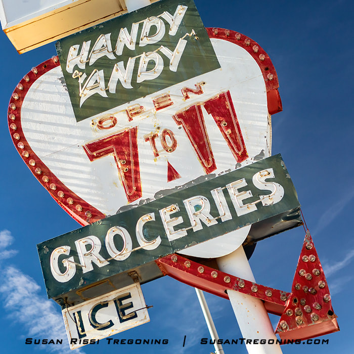 The Handy Andy Groceries neon sign stands on a tall metal pole beside the roadway. The vintage sign features bold lettering and a classic mid‑century design, with the sky visible behind it.