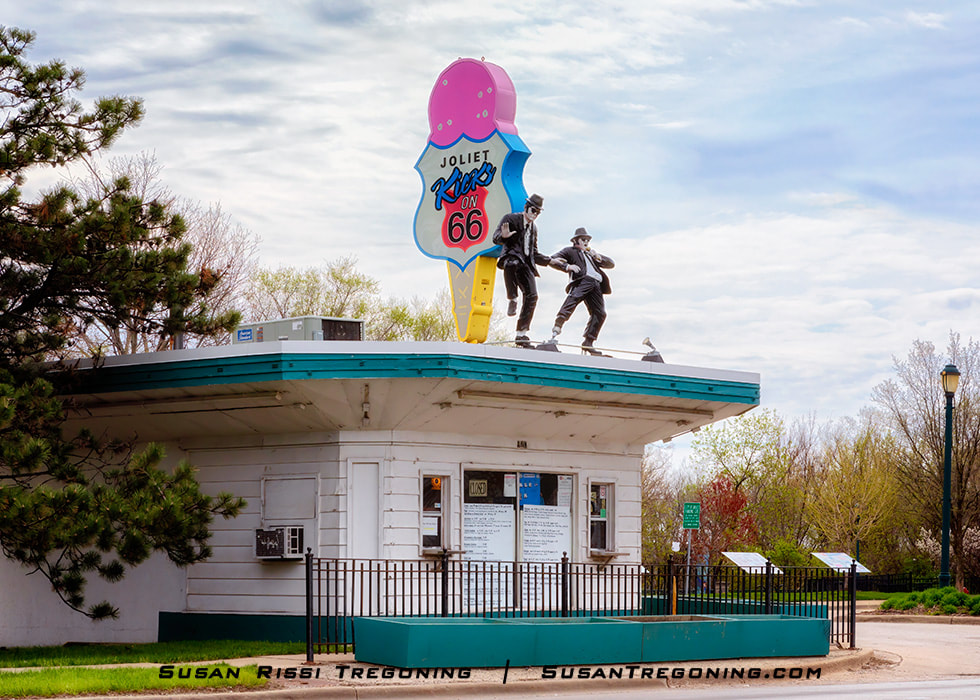 A small roadside building with a teal roof stands among trees and fencing. Two statues resembling the Blues Brothers, dressed in black suits, hats, and sunglasses, appear in dancing poses atop the roof. Above them is a large ice‑cream‑cone sign reading Kicks on 66. The structure is part of Route 66 Park in Joliet, Illinois.