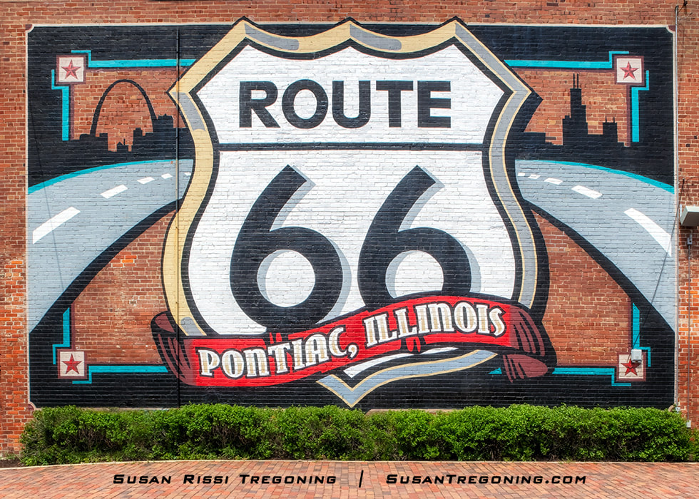 A large mural painted on a brick wall features a Route 66 shield above a red banner reading Pontiac, Illinois. A curved road graphic leads toward stylized skylines on each side. Red bricks cover the ground in front of the mural; these are original Route 66 bricks designed to be driven on. The mural sits behind the Illinois Route 66 Hall of Fame Museum Complex.