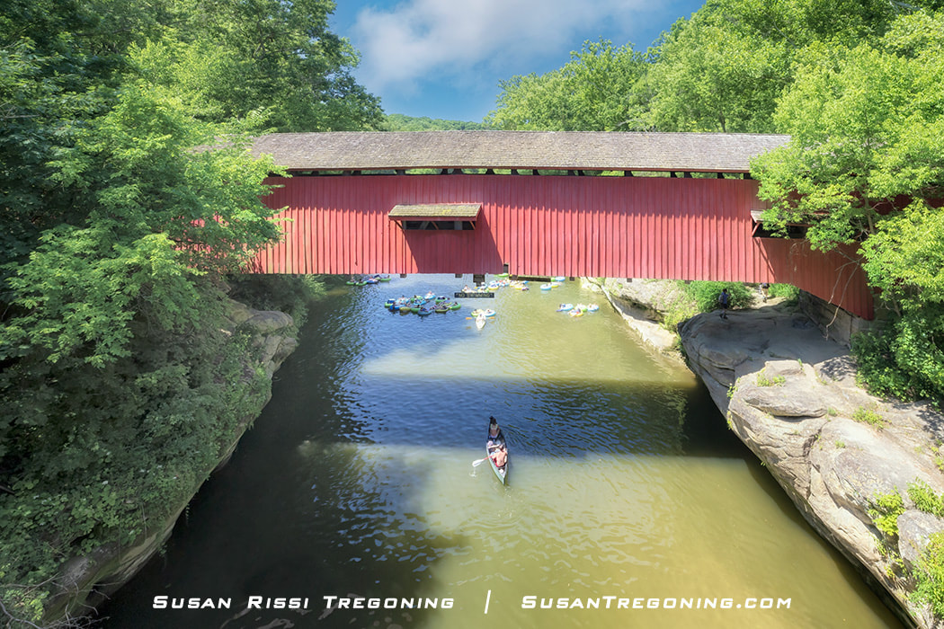   
People float down Sugar Creek on inner tubes and paddle a canoe beneath the red Narrows Covered Bridge, surrounded by lush green trees and calm summer water.