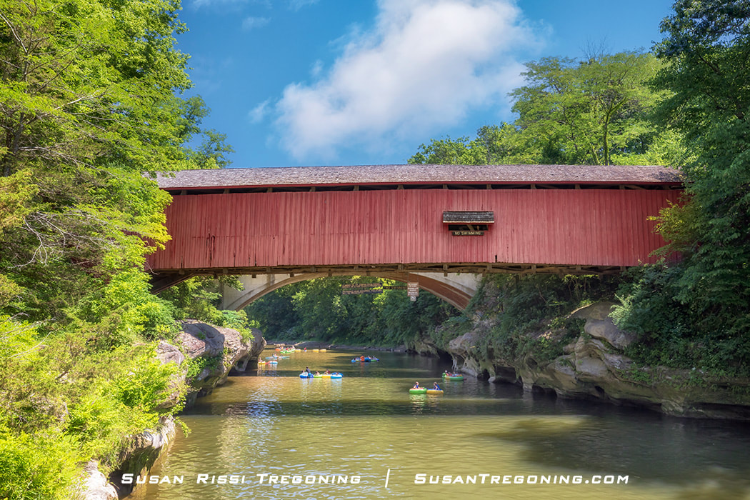   
People float down Sugar Creek in kayaks and inner tubes beneath the red Narrows Covered Bridge, surrounded by lush green trees and calm summer water.