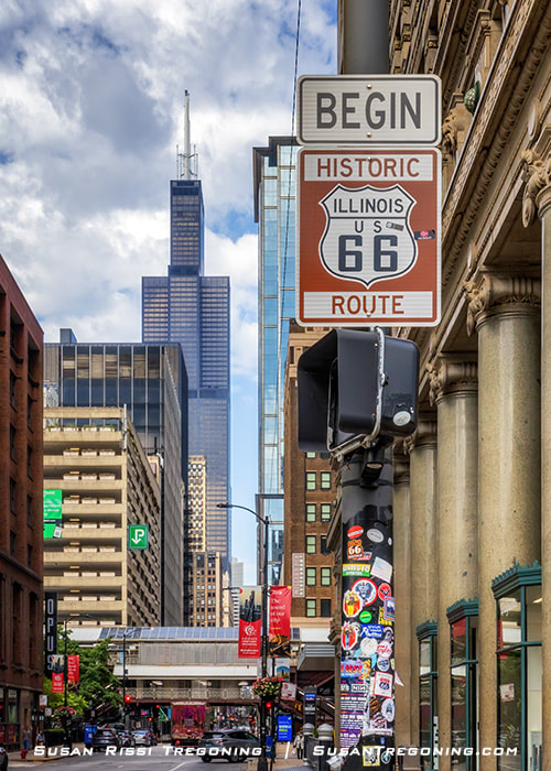 Route 66 Begin sign at Michigan Avenue and Adams Street in Chicago, with tall downtown buildings and the Willis Tower visible in the background.