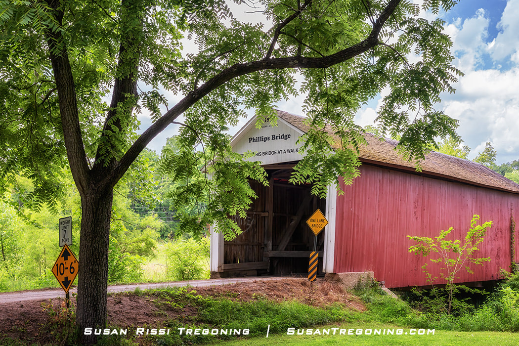   
#22 Phillips Covered Bridge, a red wooden bridge with a white gabled entrance, set along a dirt road and surrounded by lush green trees, with posted clearance and weight‑limit signs near the entrance.
