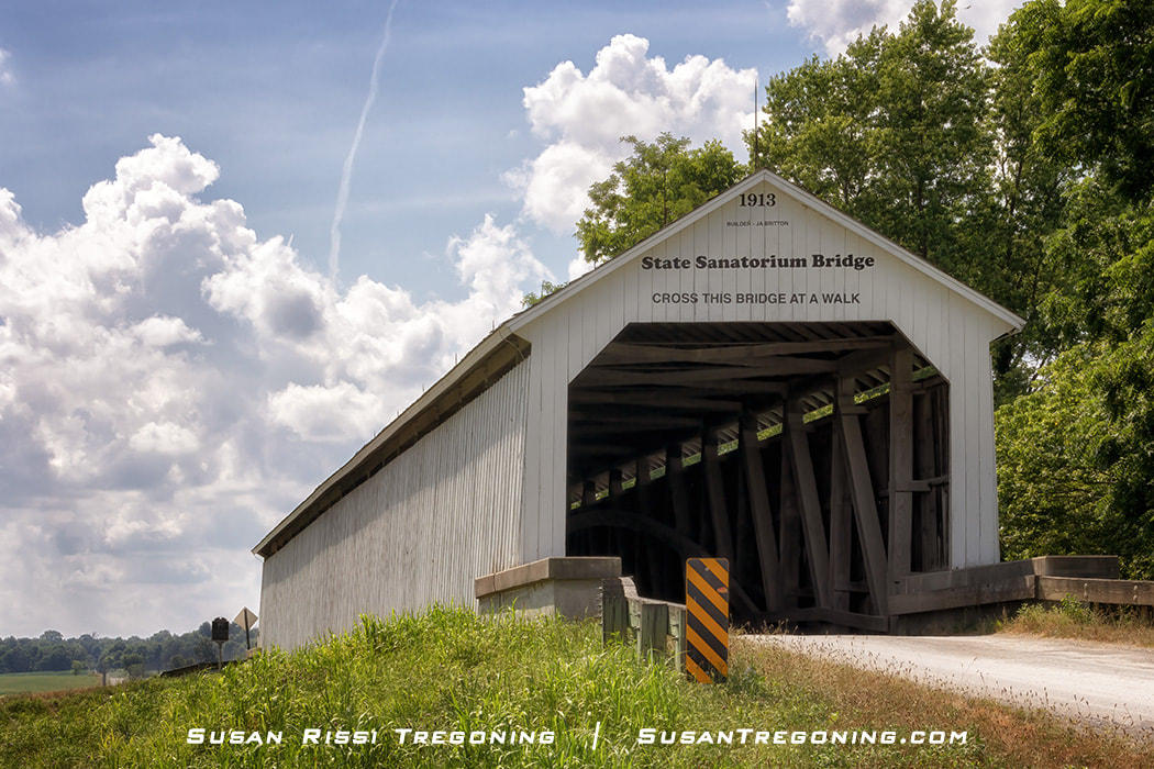 White-painted covered bridge known as the State Sanatorium Covered Bridge, relocated to its current public site in 2008. The bridge stands over a small waterway with trees and natural surroundings visible.