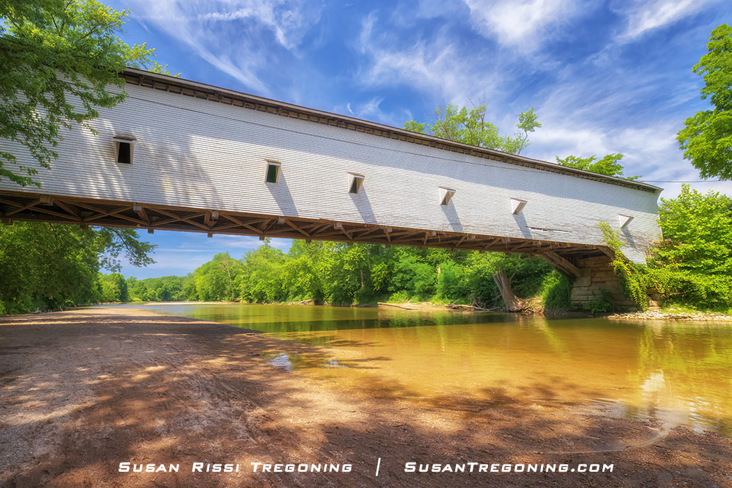 The Jackson Covered Bridge in profile, a long white wooden span with small rectangular side windows, crossing Sugar Creek and surrounded by green trees under a bright blue sky.