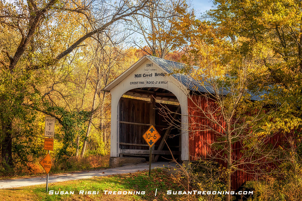 Mill Creek Covered Bridge, a white‑entrance, red‑sided wooden span dated 1907, set along a gravel road and surrounded by yellow and orange autumn foliage with posted weight‑limit and clearance signs.