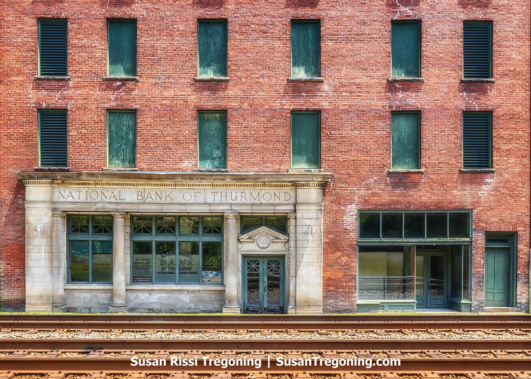 Front view of the historic National Bank of Thurmond building in West Virginia, a brick structure with a stone‑framed entrance and boarded windows, positioned directly beside railroad tracks in the narrow downtown corridor.
