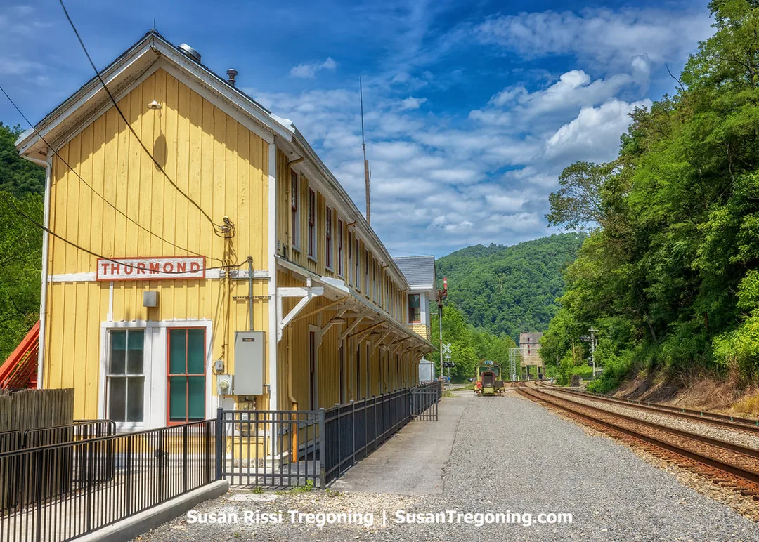 The two‑story yellow Thurmond Train Station in West Virginia, a 1904 wooden depot with white trim and a slanted first‑floor platform roof, stands beside railroad tracks that curve into a forested Appalachian valley under a blue sky with scattered clouds.