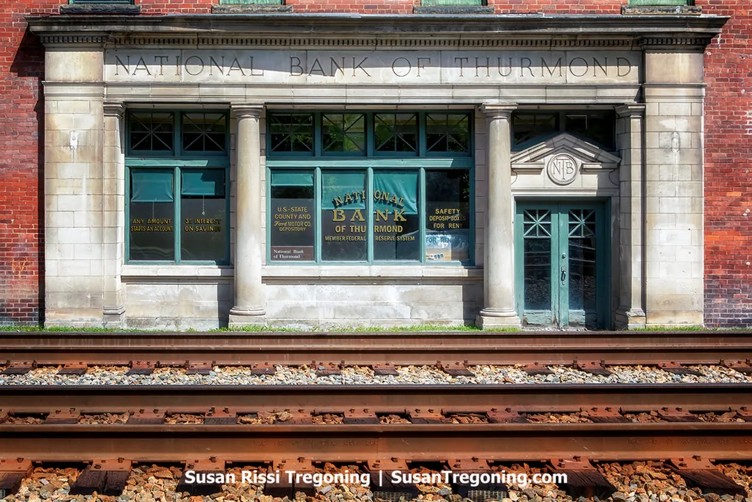 Front view of the historic National Bank of Thurmond building in West Virginia, a brick structure with stone detailing and large green‑framed windows, positioned beside active railroad tracks.