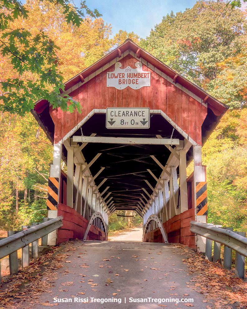   
A red covered bridge with white trim stands at the entrance to a forested roadway lined with autumn foliage in shades of orange, yellow, and green. A sign above the portal reads 1891 Lower Humbert Bridge, with a clearance sign below it. The wooden interior truss structure is visible through the opening. 
