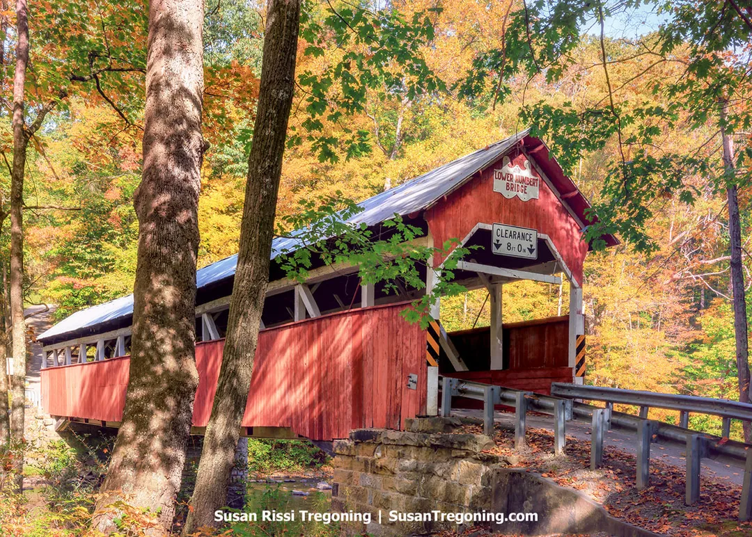 A red covered bridge with white trim spans a small stream, surrounded by autumn trees in shades of yellow, orange, and green. A sign above the entrance reads Lower Humbert Bridge with a posted clearance of 8 feet 0 inches. Stone abutments and metal guardrails appear in the foreground.