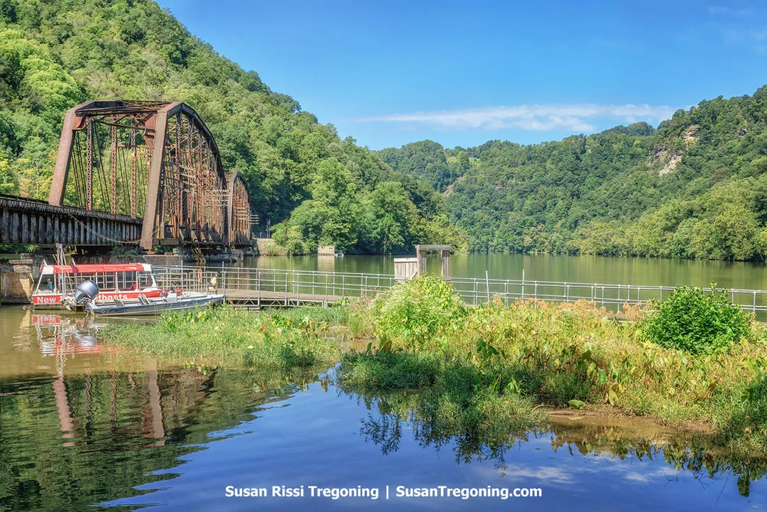 A rust‑colored truss bridge spans the river between forested hills, with a red and white jet boat docked in the foreground beside shallow water and aquatic plants under a clear blue sky.
