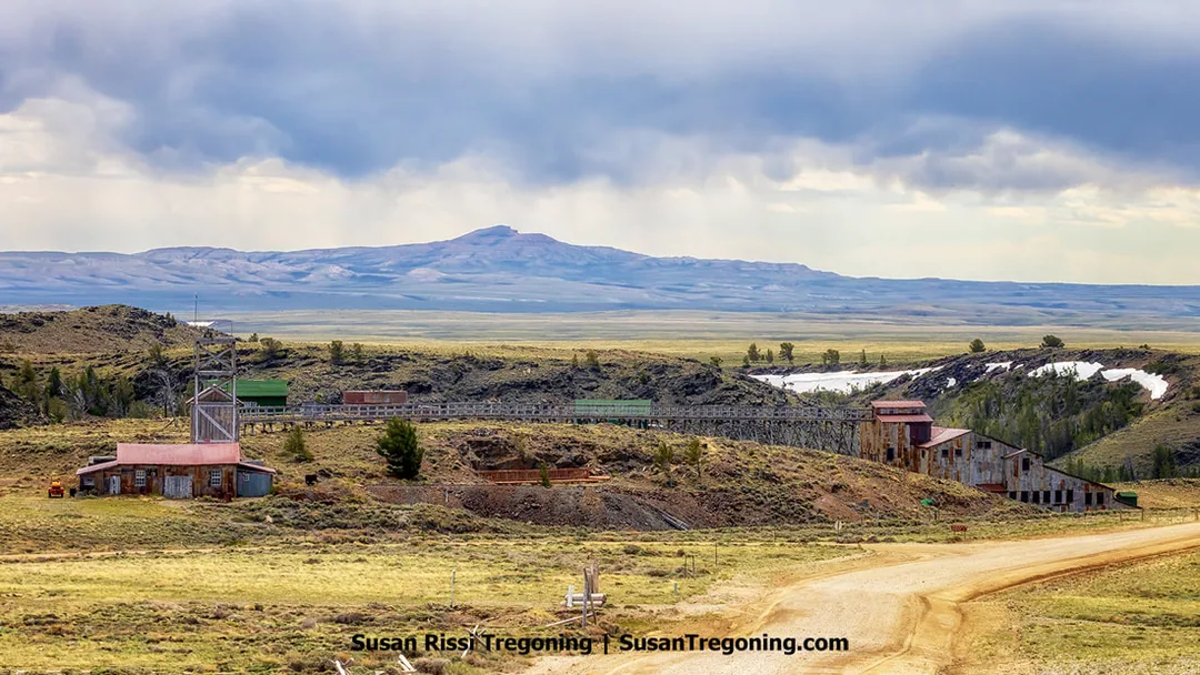 Wide view of the Carissa Mine and Mill at South Pass City, showing weathered wooden and metal buildings, a tall framework tower, and a long elevated trestle on open grassland. Dark clouds hang over the surrounding mountains.
