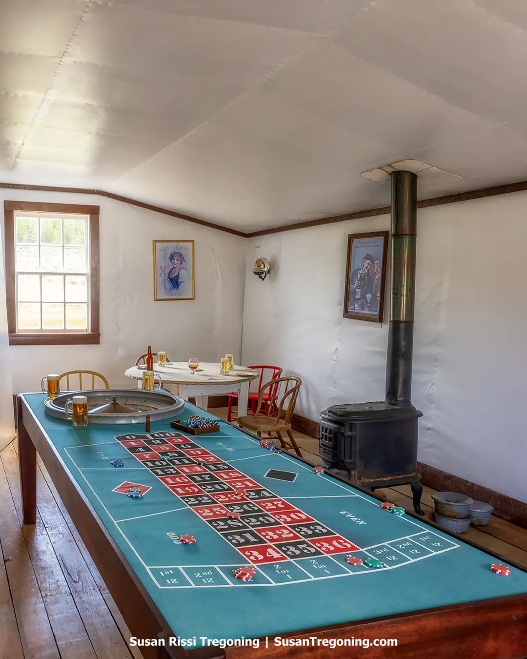 Interior of a historic gaming room with a roulette table covered in teal felt, showing numbered betting squares, poker chips, and a roulette wheel. A round table with chairs and glasses sits in the background. A black wood‑burning stove stands near the wall, with framed portraits and a window providing natural light.