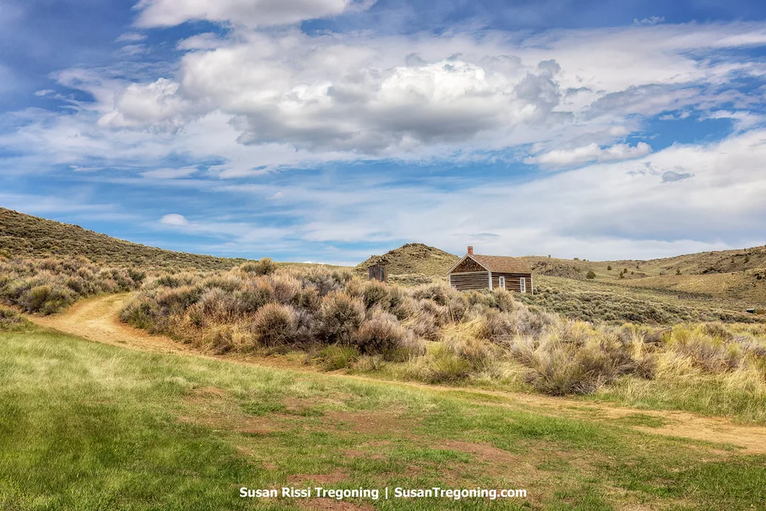 Log one‑room schoolhouse perched on a hill above South Pass City, with sagebrush and open grassy slopes in the foreground and a partly cloudy sky overhead.