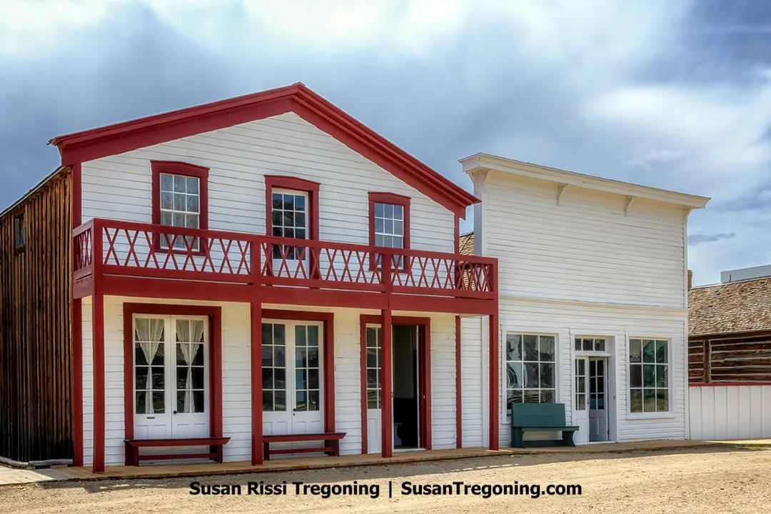 Two preserved nineteenth‑century wooden buildings in South Pass City, including the South Pass Hotel and Restaurant, with white siding, red trim, upper‑story balconies, and large front windows facing a dirt street beneath a partly cloudy sky.
