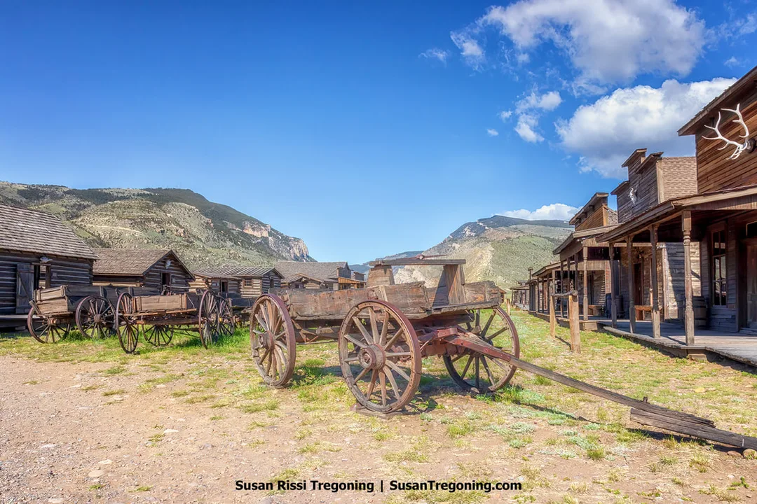A row of wooden wagons with large spoked wheels positioned between rustic wooden buildings in a historic Old West town setting, with mountains and a partly cloudy sky in the background.
