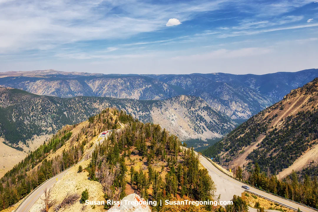     An elevated view above a winding mountain road with rugged cliffs, forested slopes, and deep valleys extending into the distance beneath a partly cloudy sky.

