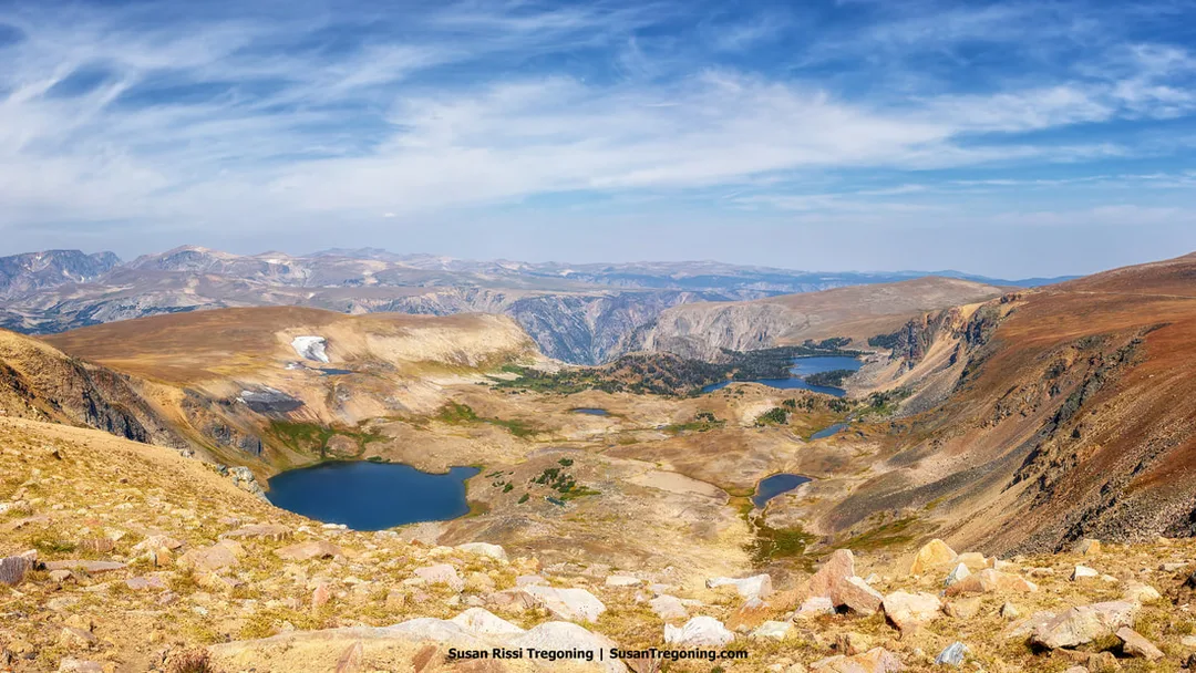 A high‑elevation mountain landscape with rocky slopes, sparse alpine vegetation, and several small lakes across a broad valley. Steep cliffs and distant ridges rise beneath a partly cloudy sky.