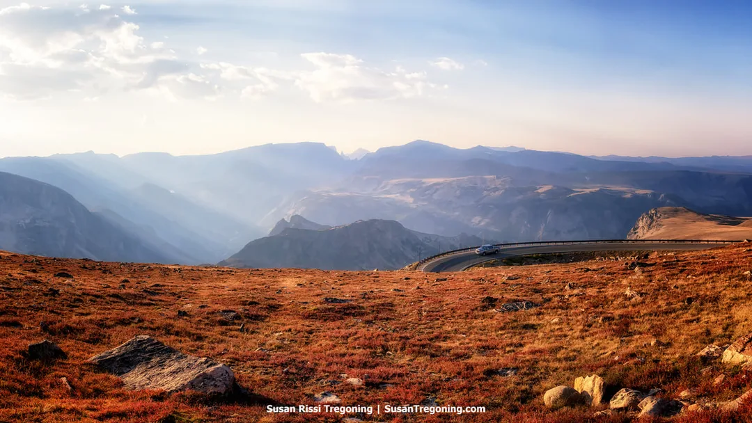 A high‑elevation view along the Beartooth Highway shows a paved road curving across an open alpine plateau. Reddish‑brown tundra and scattered rocks fill the foreground, with deep valleys and layered mountain ridges extending into the distance. Sunlight filters through clouds, creating soft rays across the scene. A single vehicle travels along the road, emphasizing the scale of the surrounding landscape.