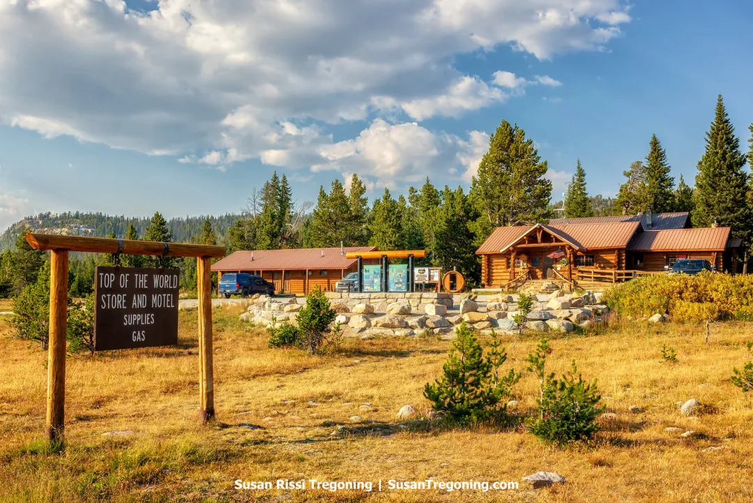 A rustic log building complex surrounded by pine trees and distant mountains beneath a partly cloudy sky. A wooden roadside sign stands in the foreground, and several vehicles and gas pumps are visible near the buildings.