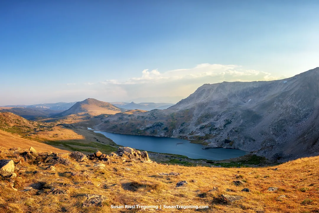  A high‑elevation landscape with a lake nestled between rocky slopes and grassy terrain. The foreground shows golden‑brown grass and scattered rocks, with distant mountain ridges rising beneath a mostly clear sky.