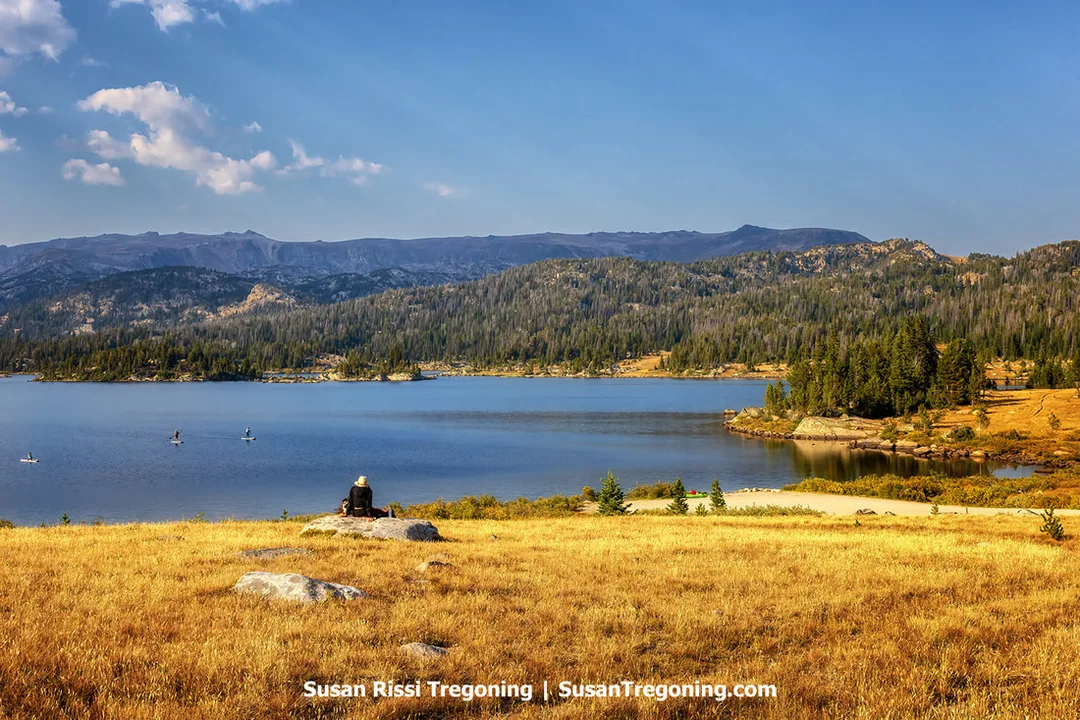A large mountain lake bordered by forested hills and distant ridges beneath a mostly clear sky. The foreground shows golden grass and scattered rocks, with a person seated on a rock near the shoreline and several small boats visible on the water.