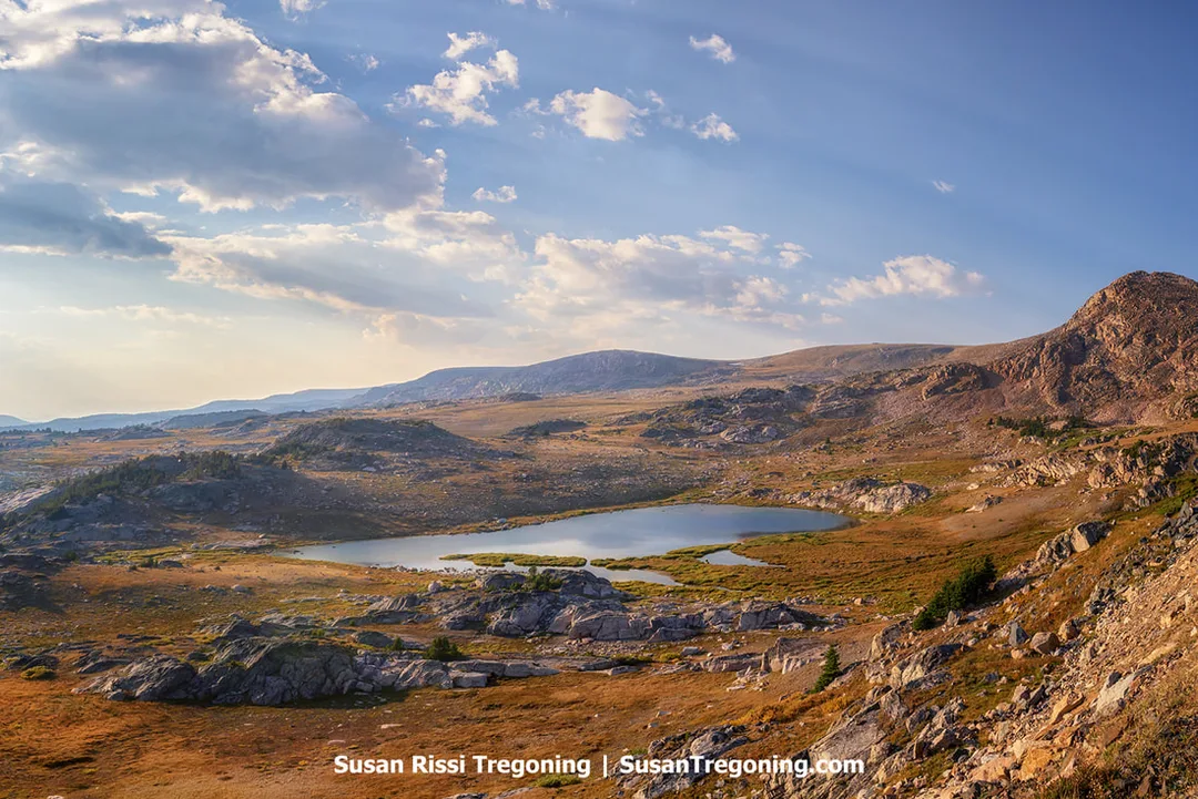 A high‑elevation alpine landscape with a small lake surrounded by rocky ground and open grassy areas. Rolling hills and distant mountain ridges extend toward the horizon beneath a partly cloudy sky.