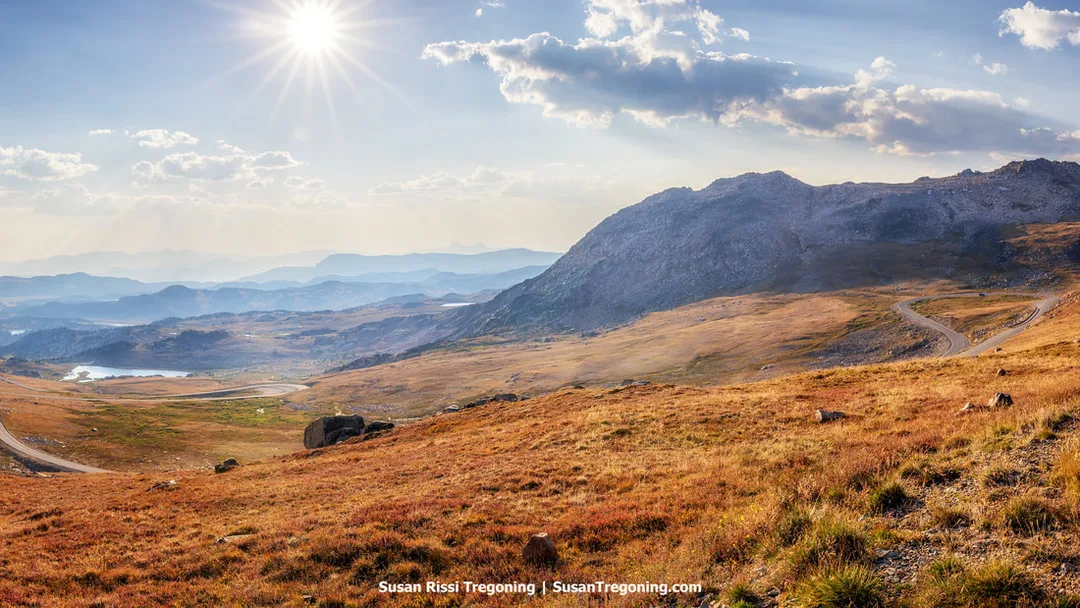A high‑elevation landscape above the tree line with golden‑brown tundra, scattered rocks, and a winding paved road. A rugged mountain rises to the right, with distant hazy ridges and a small lake visible beneath a partly cloudy sky and bright sun.