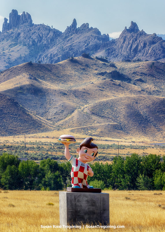 A Big Boy statue stands on a concrete pedestal in an open field of golden grass. Behind it, scattered trees and distant buildings sit at the base of rugged, jagged mountains rising sharply under a clear blue sky.