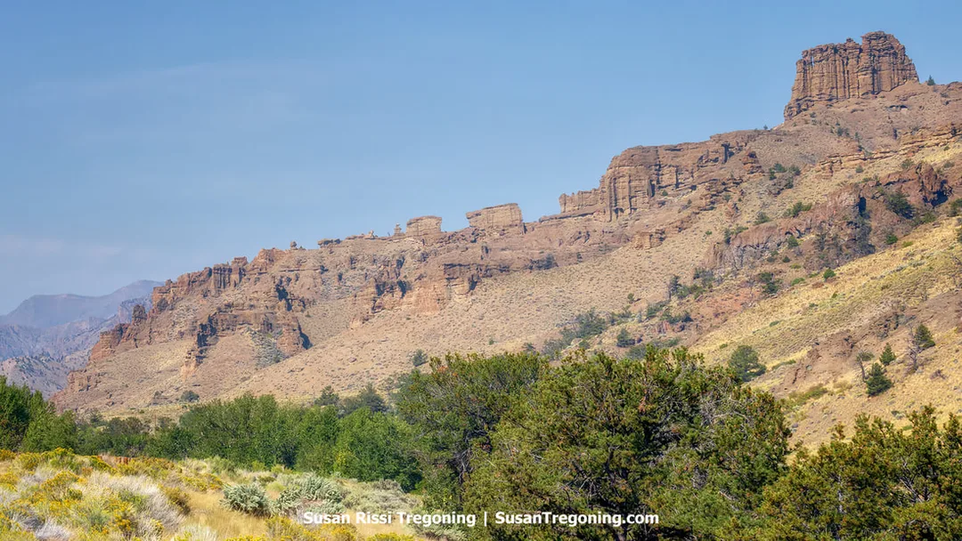 Steep, rugged cliffs with layered rock formations rise across a dry mountainside, with sparse shrubs and small trees scattered along the slopes and valley floor. The eroded strata and jagged ridges are clearly visible beneath a bright, clear blue sky.