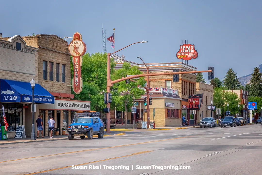 A view of downtown Cody, Wyoming, showing early twentieth‑century commercial buildings with brick and stone façades, business signs, parked vehicles, and people walking along the sidewalk under a clear sky.