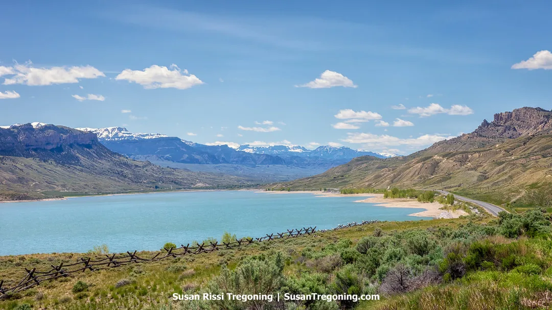   
A large turquoise reservoir sits below rolling hills and rugged mountains, with snow‑capped peaks in the distance. A wooden fence and green vegetation line the foreground, and a road curves along the shoreline on the right under a bright blue sky with scattered clouds.