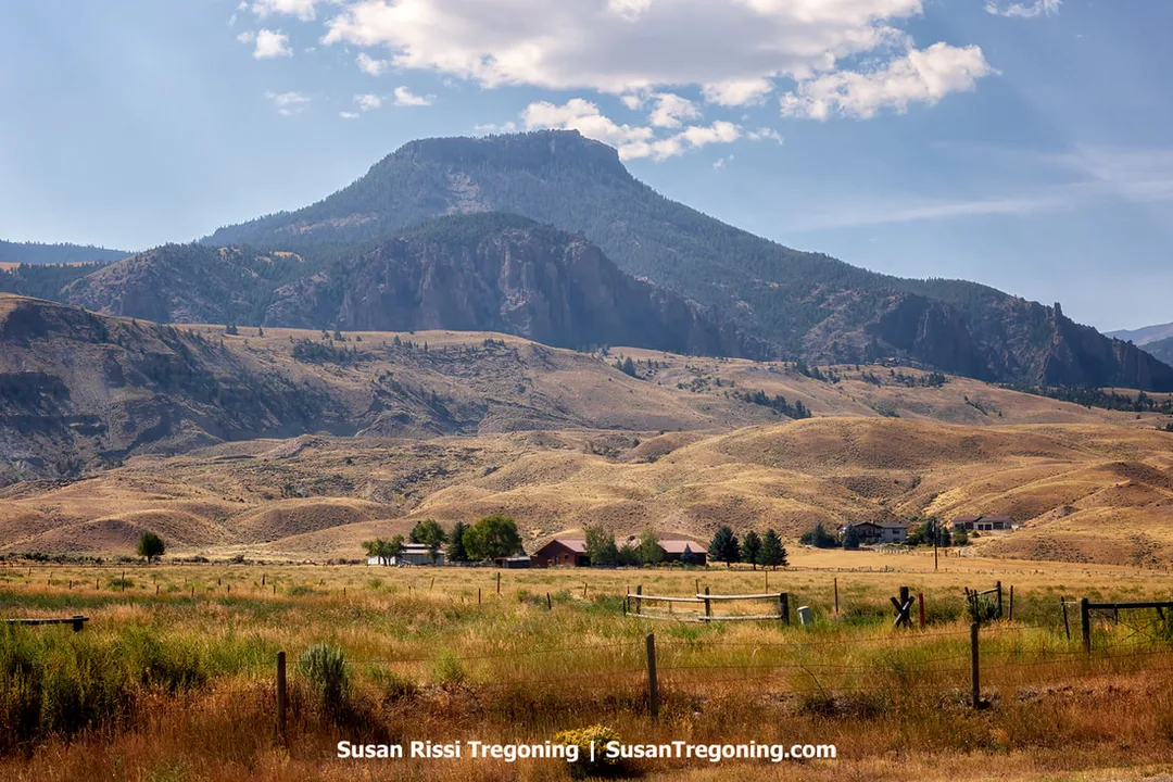 A fenced field with green and yellow grass sits in the foreground, with several houses and barns surrounded by trees in the mid‑ground. Behind them, a large rugged mountain rises in steep, rocky layers under a partly cloudy sky.