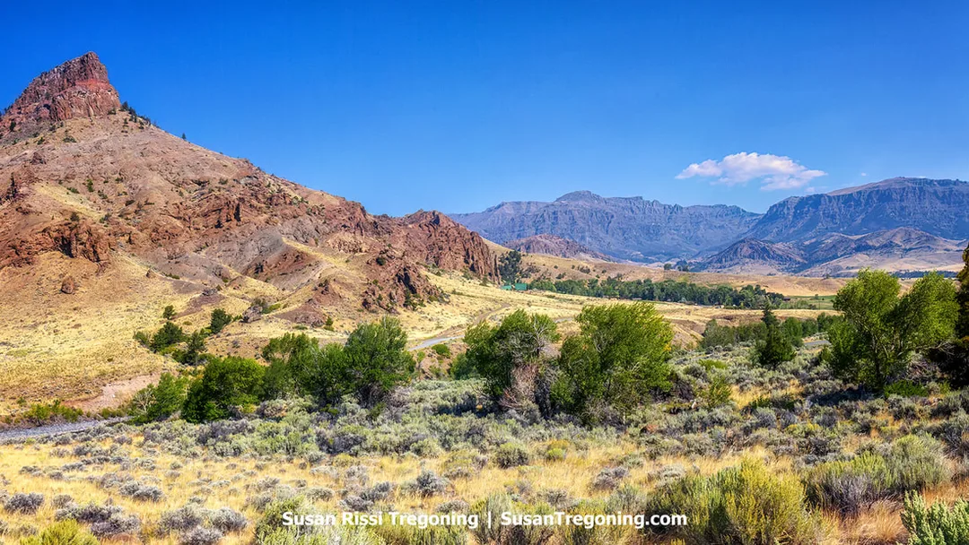 Reddish‑brown rocky hills rise in the foreground with dry grass, scattered shrubs, and clusters of green trees. Behind them, a range of blue‑gray mountains stretches across the horizon under a clear blue sky.