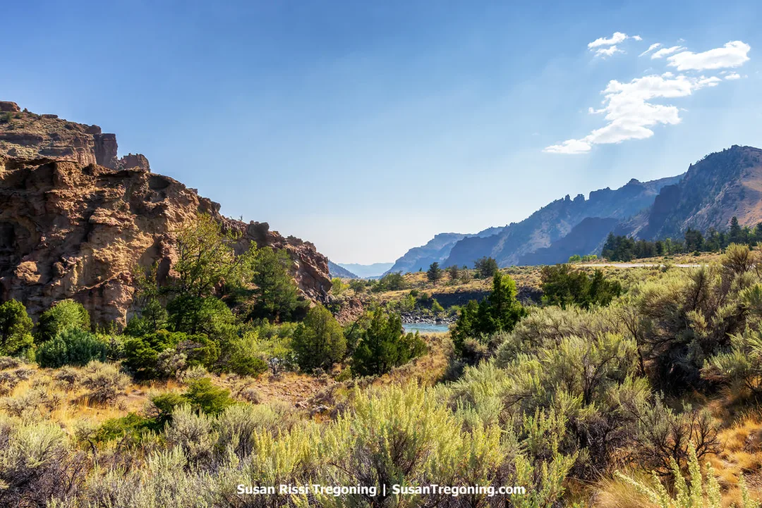 Rugged rock formations rise on the left above dense shrubs and small trees, with a river flowing through the middle ground. Distant mountain ranges appear beyond the valley under a clear blue sky with a few scattered clouds.