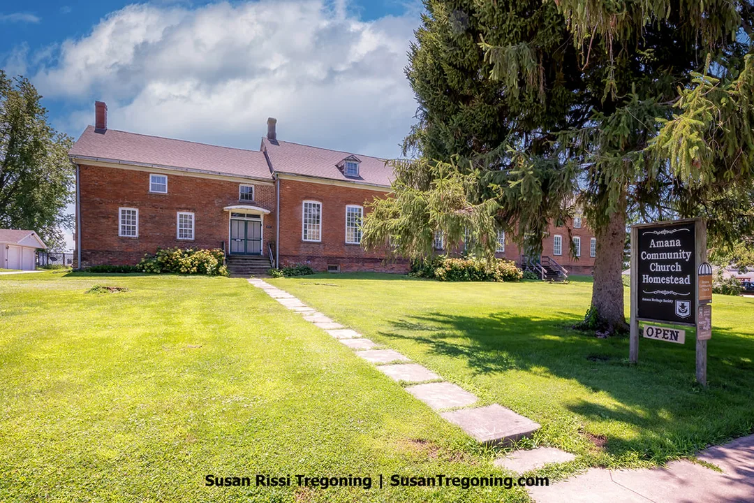 The exterior of the Amana Community Church Museum in Homestead, Iowa, showing the preserved historic brick church with its simple façade and visible windows. The building sits within the village setting of the Amana Colonies.