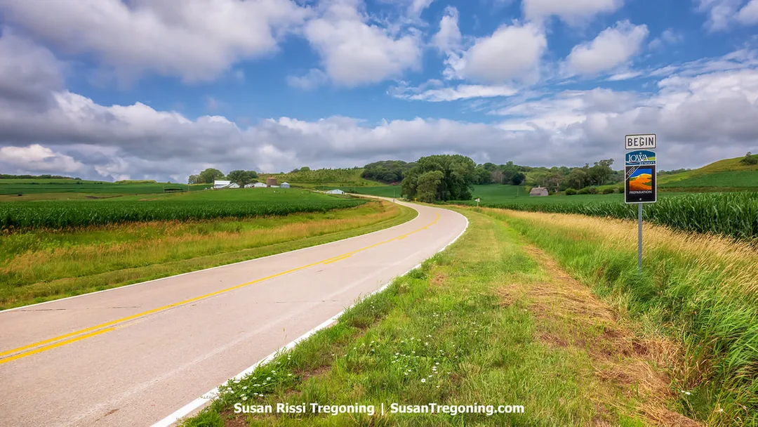 A roadside perspective at the beginning of the Preparation Loop along Iowa’s Loess Hills National Scenic Byway. A byway sign stands near the road, with green cornfields and the region’s rolling, loess‑formed hills extending into the distance.