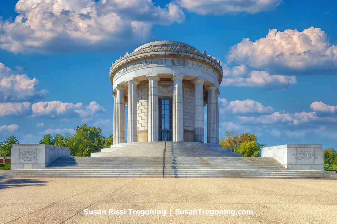 The circular, columned George Rogers Clark Memorial stands at George Rogers Clark National Historical Park in Vincennes, Indiana. The neoclassical structure features a domed roof and tall stone columns, with engraved inscriptions on nearby stone blocks. Trees frame the scene under a partly cloudy sky.