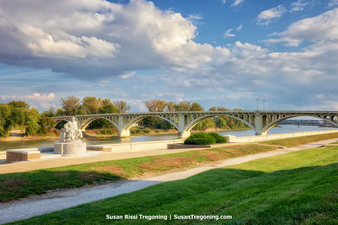 A fully stone statue of Francis Vigo sits on a stone pedestal at George Rogers Clark National Historical Park in Vincennes, Indiana. The statue overlooks the Wabash River, where the Lincoln Memorial Bridge crosses the water in a series of concrete arches. Trees line the far bank beneath a partly cloudy sky.