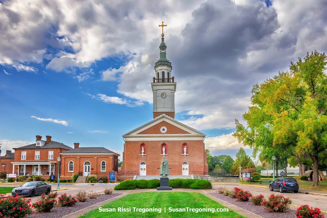 The historic Basilica of St. Francis Xavier, part of the Old Cathedral Complex in Vincennes, Indiana, stands along a town street. The brick church features a tall steeple topped by a gold cross, with three arched façade niches holding statues and a larger statue on a pedestal in the landscaped garden at the entrance. Nearby brick buildings and parked cars line the street beneath a partly cloudy sky.