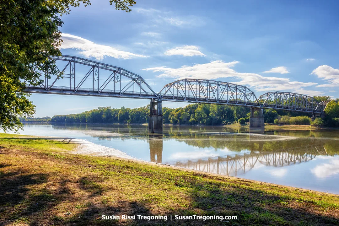 The abandoned Harmony Way Toll Bridge spans the Wabash River in a long steel truss, its repeating triangular framework reflected clearly in the calm water below. Concrete piers rise from the river to support the structure, and tree branches frame the view from a grassy, sandy riverbank in the foreground. A backdrop of wooded shoreline and a bright sky gives the scene a quiet, peaceful atmosphere despite the bridge’s disuse.