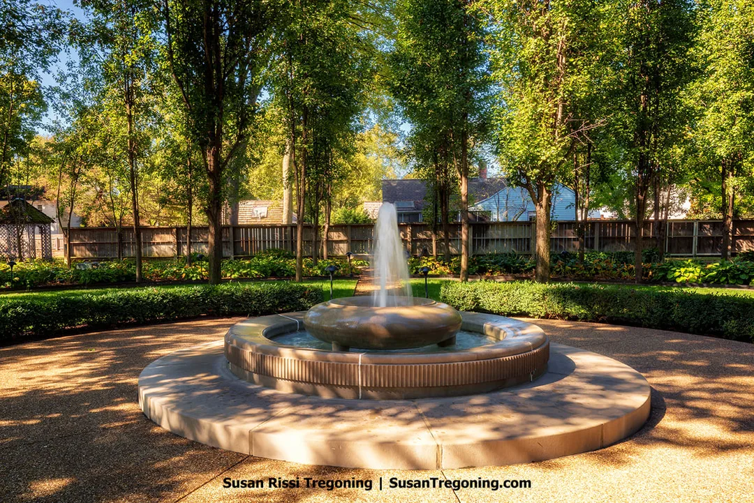 A circular stone fountain sprays water upward from a raised central basin, set within a paved courtyard bordered by neatly trimmed hedges. Tall trees cast dappled shade around the Fountain of Life, with a wooden fence and glimpses of nearby homes visible through the foliage, creating a quiet, residential garden setting.