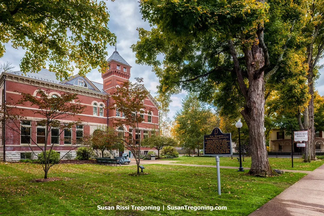 A tall red‑brick building with a central tower stands behind a lawn lined with benches, lampposts, and mature autumn trees. A historical marker sits near the walkway, and the structure’s arched windows and decorative brickwork give New Harmony’s Working Men’s Institute a stately civic presence within the park‑like setting.