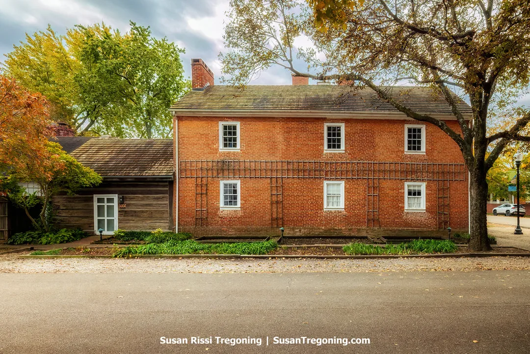 A two‑part historic building combines an early log structure on the left, built of horizontal timbers with white chinking and a simple door and window, with a two‑story red‑brick section on the right featuring white‑trimmed windows and a gabled roof. A wooden trellis leans against the brick façade, and autumn trees rise behind the building. A landscaped garden bed borders the front, and a paved street and lamppost frame the scene.