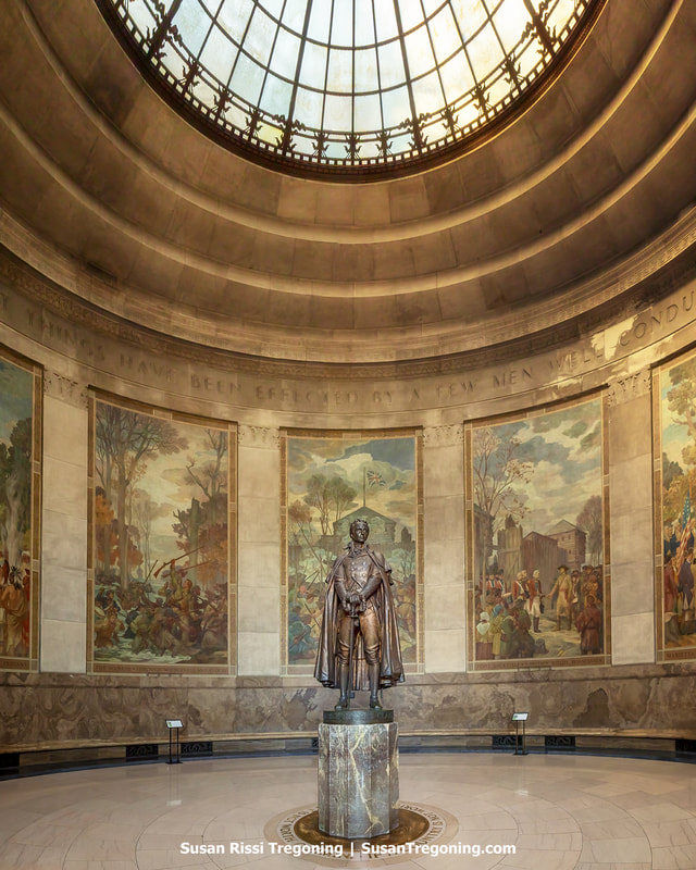 Inside the domed rotunda at George Rogers Clark National Historical Park, a statue of George Rogers Clark stands on a marble pedestal beneath a large circular skylight. Surrounding the statue are expansive historical murals framed by stone columns, with an inscription above them reading, “Great things have been effected by a few men well conducted.” Soft light from the skylight illuminates the space.