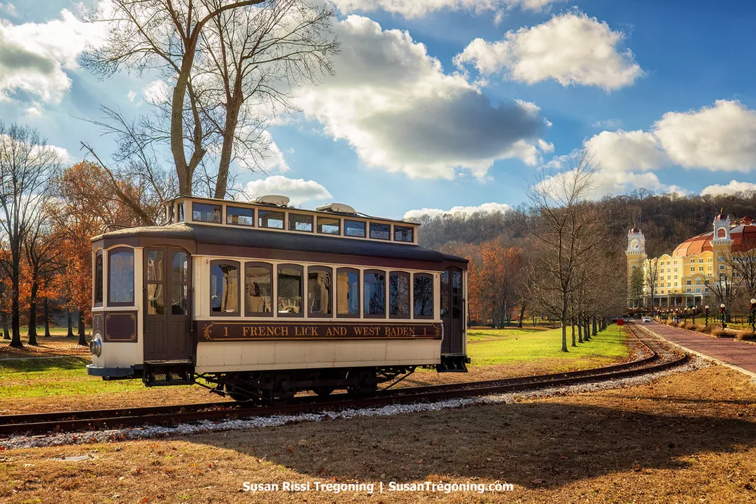 A vintage trolley labeled “French Lick and West Baden Railway” travels along a curved track through a park-like setting. Leafless trees line the path, and a large yellow historic hotel with red roofs and towers rises in the background under partly cloudy sunlight.