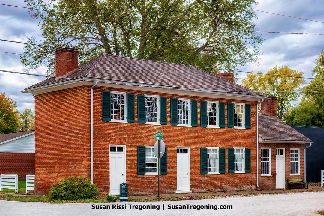A two‑story red‑brick house with green shutters stands at a street corner, its white doors and paired chimneys giving it a simple early‑nineteenth‑century appearance. A street sign marks the intersection beside the building, and autumn trees with yellow and orange leaves rise behind a white fence, adding seasonal color to the historic 1820 Owen House.